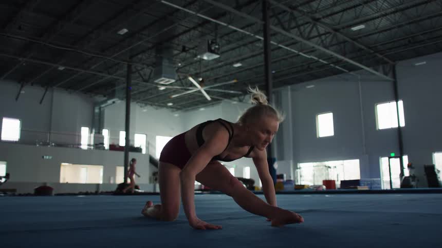 Inside a spacious gymnasium a focused female gymnast wearing a sports bra and shorts performs flexibility training on a blue mat transitioning from a kneeling lunge into a deep forward hamstring stretch