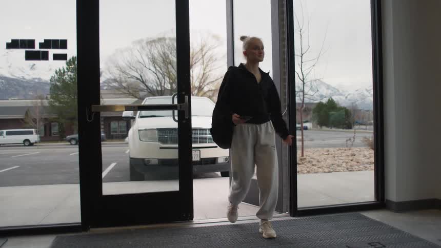 A young woman with her hair pulled into a high bun dressed in comfortable athletic wear and carrying a black backpack opens a set of glass doors to walk from an outdoor parking lot into a building lobby