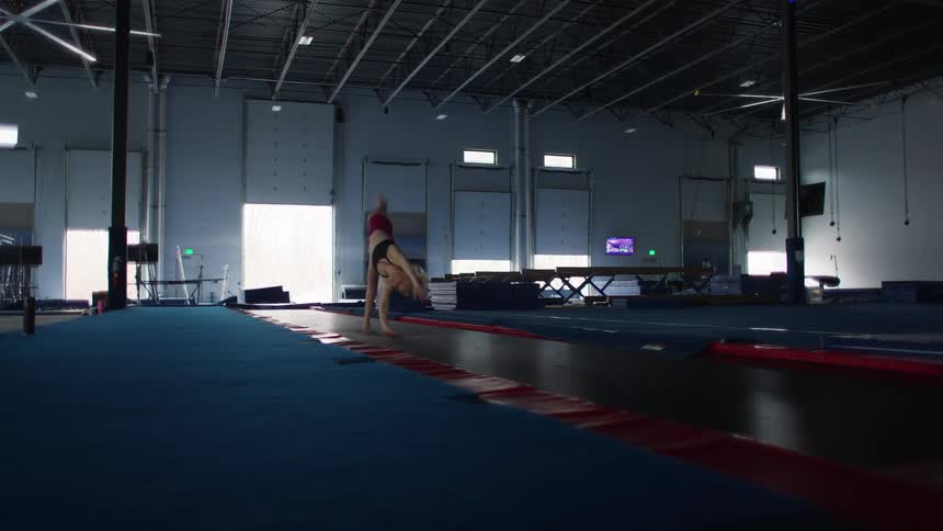 She begins her runup on a blue mat executes a roundoff into a back handspring on a red and black tumbling track and finishes with a high arcing back tuck or layoutthe gym features white pillars large rollup doors and various gymnastics apparatus