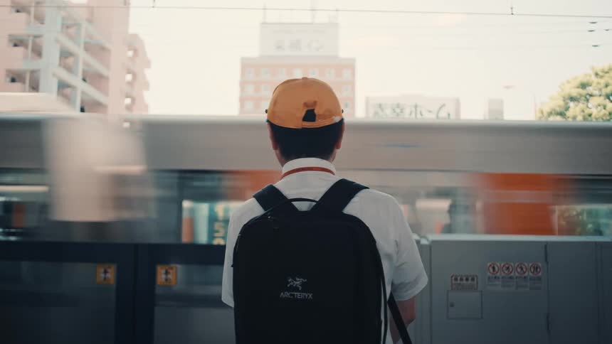 A back view of a businessman wearing a white shirt and an orange baseball cap with a black backpack standing on a train platform as a blurred train passes quickly in front of him