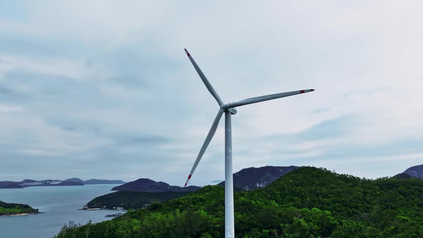 A single wind turbine with white blades stands tall on a lush green mountainous landscape and a lake against a cloudy blue sky