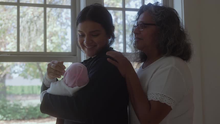 A medium shot shows a young woman holding a newborn baby wrapped in a pink cap while standing by a large window as an older woman with glasses places her hand on the young womans shoulder smiling