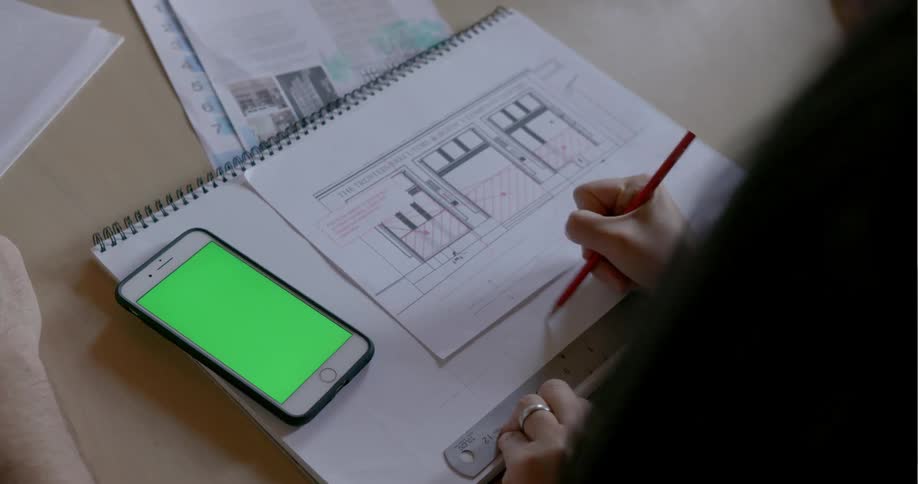 A woman sits at a table drawing with a pencil and a ruler in a sketchbook