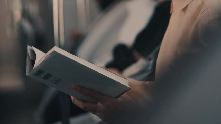 A persons hands holding an open paperback book with a plain white cover actively engaged in reading while sitting inside what appears to be a moving public transportation vehicle