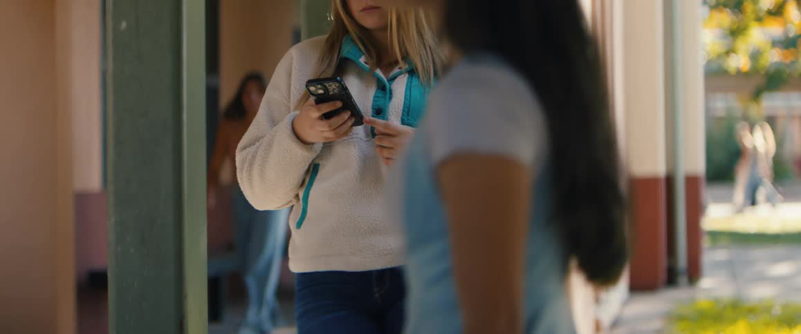 A medium shot of a young woman standing outdoors in a beige fleece pullover with a teal collar scrolling on her black smartphone as people walk past in the background