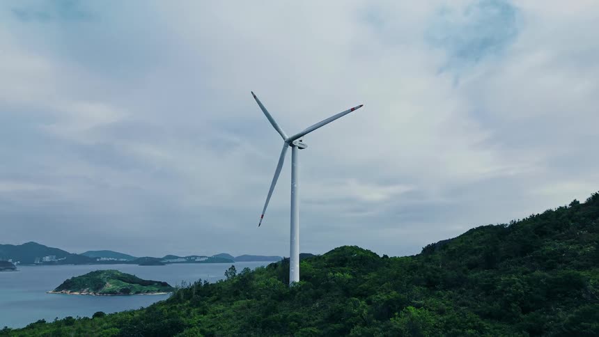 A single wind turbine stands atop a lush green hill with a calm sea and distant islands visible under a cloudy sky