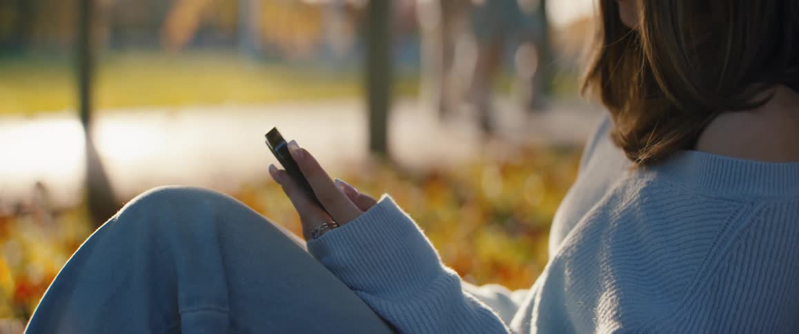 A young woman with brown hair wearing a light blue sweater and jeans sits relaxed outdoors in an autumn setting while scrolling on a smartphone