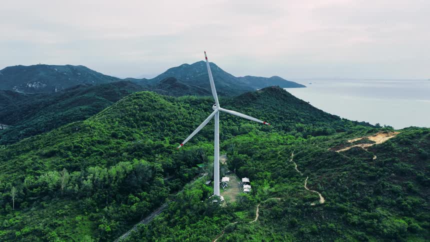 An aerial view captures a large solitary wind turbine rotating slowly on the crest of a lush heavily forested green hill surrounded by a chain of other green mountains with a sea visible in the background under an overcast sky