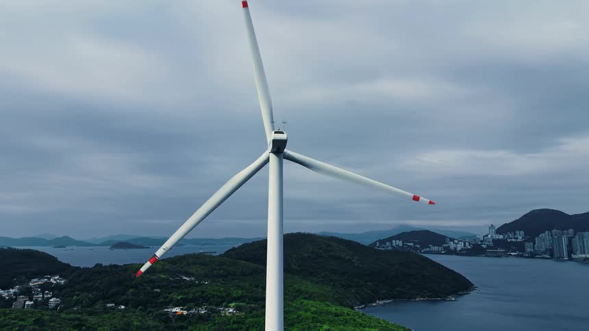 An aerial close view of a white wind turbine with red tipped blades slowly rotating against a backdrop of hilly green islands and a distant city skyline