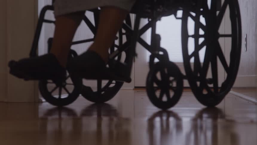 A lowangle closeup shot focuses on the wheels and lower half of a woman sitting in a black manual wheelchair as she moves forward across a reflective hardwood floor and through a white doorway