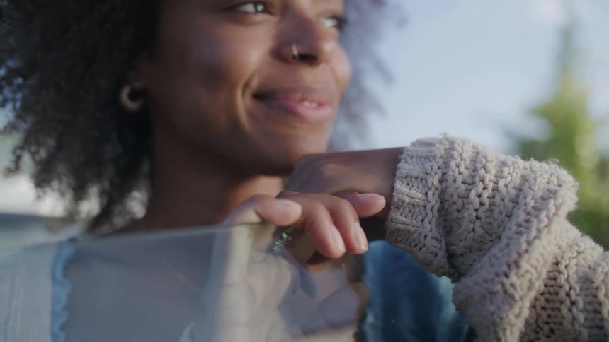 A closeup shot focuses on a young woman with curly hair who is joyfully laughing with an open mouth while resting her hand covered by a chunky knit sweater sleeve on a piece of reflective glass