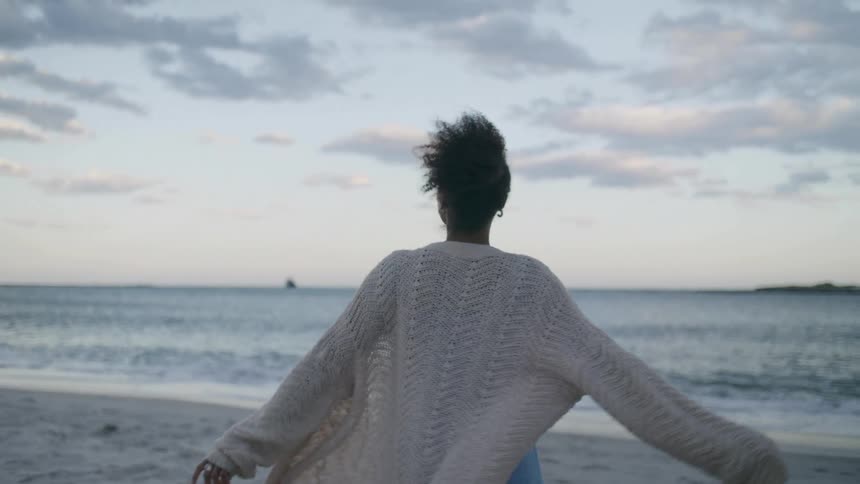 A woman wearing a light blue dress and a chunky white cardigan walks toward the water on a beach then turns to face the side and briefly smiles while spreading her arms wide enjoying the open seascape under a cloudy sky