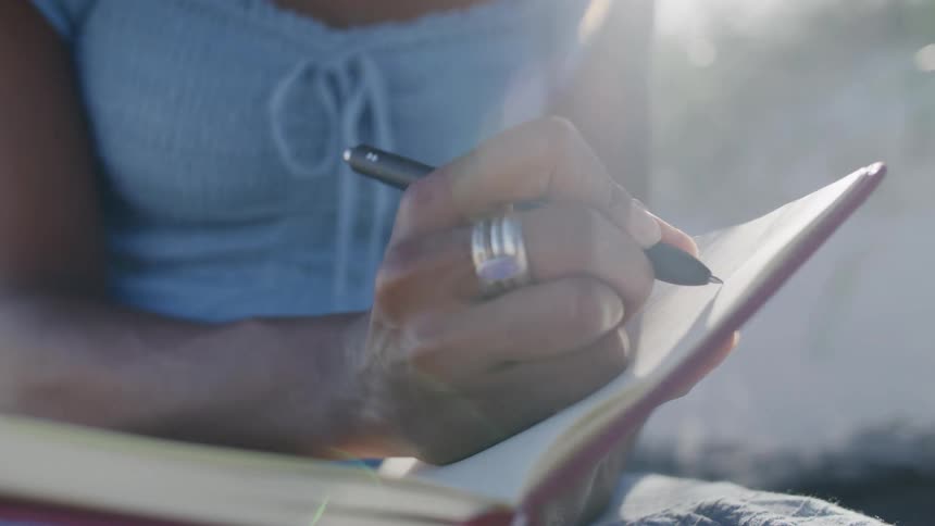 A close view shows a woman wearing a blue top and several rings writing with a black pen in a pink journal outdoors with bright sunlight flaring over her shoulder