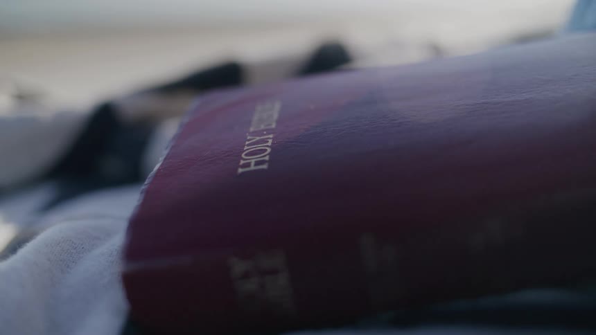 A person picks up a bible that is laying on a towel on a beach