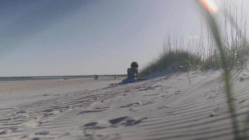Woman wearing a blue dress sitting down on the sand at a beach reading a book outdoors during day time