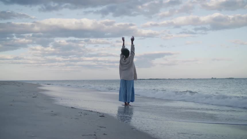 Woman wearing a sweater over a long dress stands in the water on the beach and raises her arms in the air
