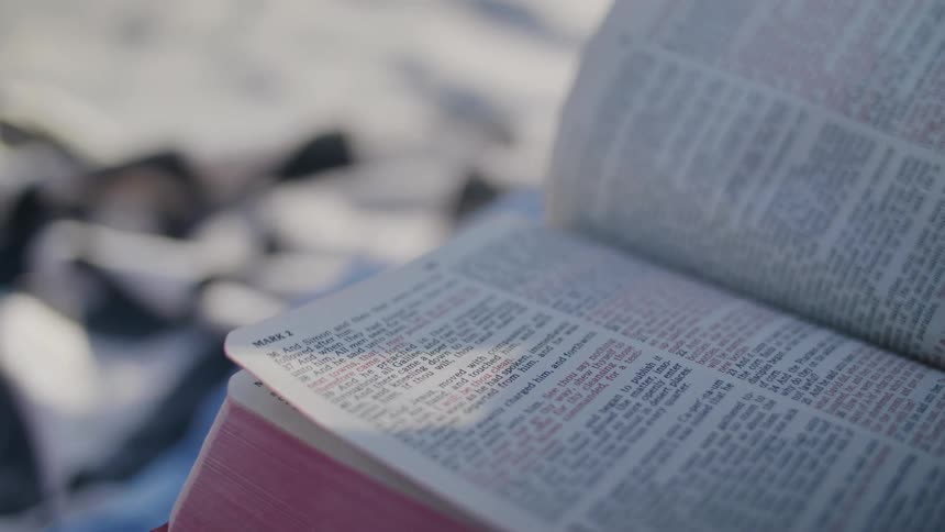 An extreme closeup shot of an open bible with the pages slightly fluttering in the breeze while resting on a blurred background object in soft natural light