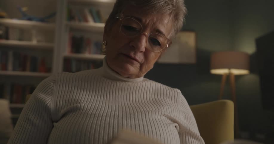 Older woman sitting at home in a room with bookcases reads out loud