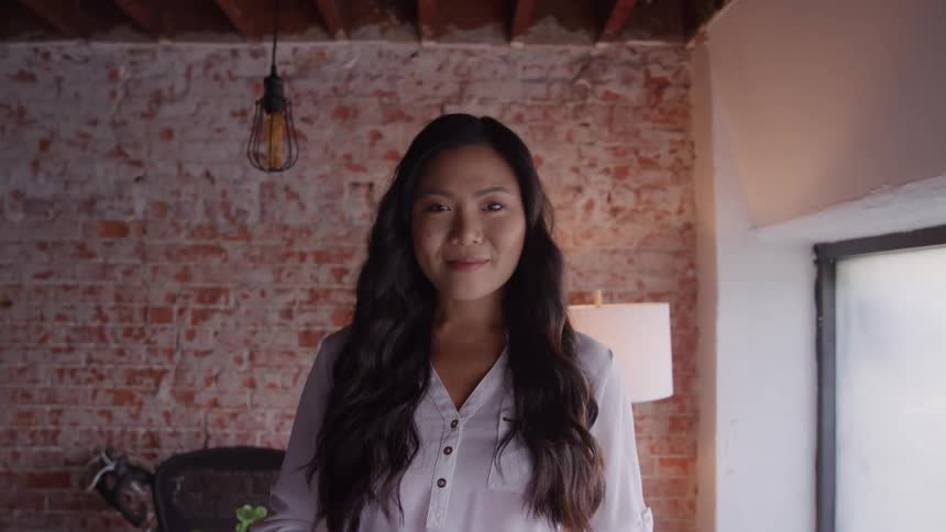 A young woman with long dark wavy hair wearing a white buttonup blouse smile for the camera standing against the background of a textured exposed brick wall in an indoor loftstyle space