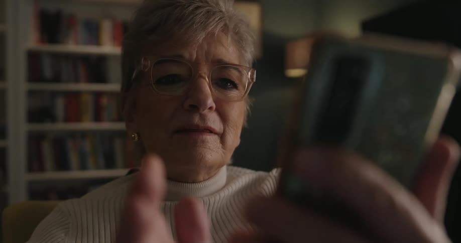 Elderly woman sits in a chair looking intently at her smartphone indoors