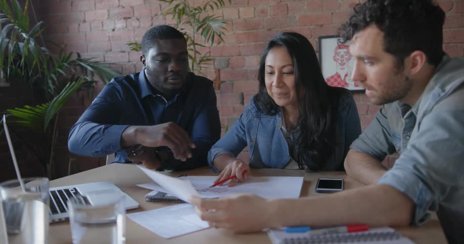 A woman and two men sit at a table looking at papers and talking