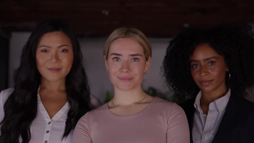 A trio of diverse professional women standing sidebyside turn their heads to face the camerathey share a confident and friendly expression standing against a dark indoor background