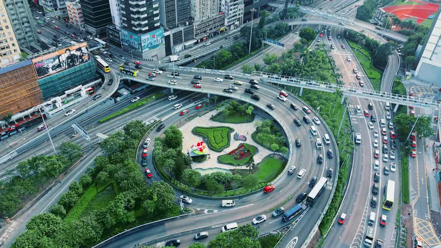 An aerial shot captures a busy complex multilevel highway interchange with continuous vehicle traffic circling a small landscaped park in the middle of a dense urban environment surrounded by tall buildings