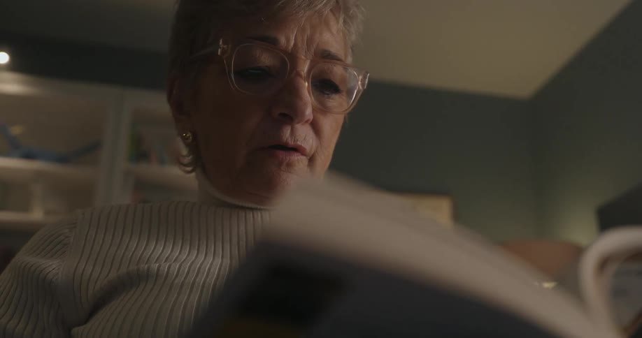 A close view of an older woman in a white ribbed turtleneck sweater and glasses reading a book aloud in a dimly lit room with a blurred white bookshelf behind her