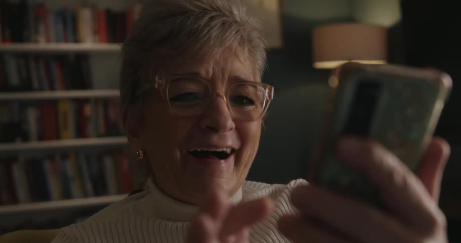 A close view of an older woman in a white turtleneck and glasses happily smiling and laughing while holding and interacting with her smartphone during a video call