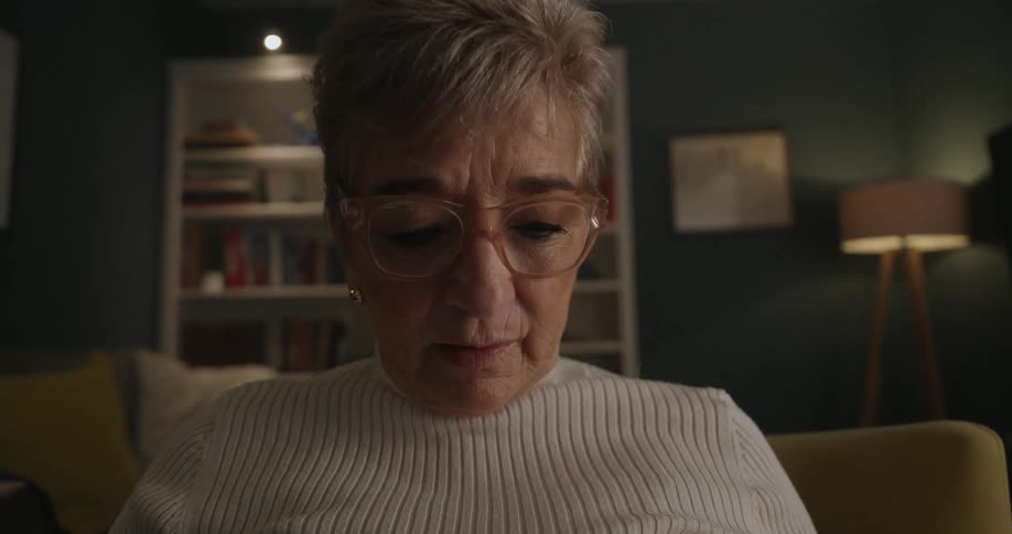Adult woman wearing glasses and a white sweater sitting down on a chair reading out loud in a living room indoors during night time