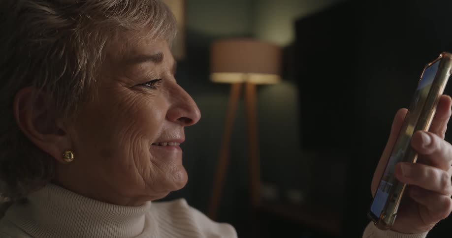 An older woman with grey hair smiles gently while holding a smartphone and looking at the screen in a living room with soft lighting