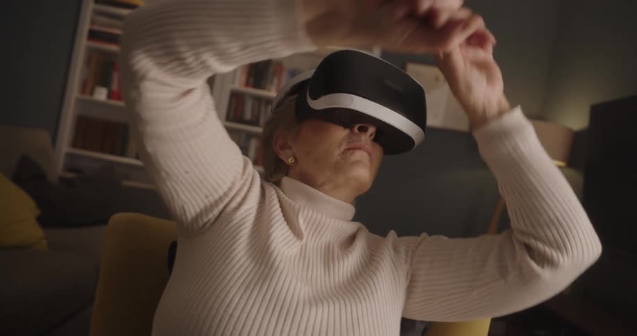 An older woman wearing a white sweater uses a virtual reality headset and moves her arms while sitting in a yellow chair in a living room