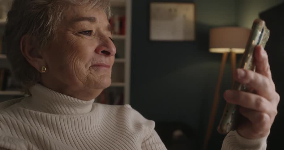An elderly woman with short blonde hair and a white turtleneck smiles and laughs while looking at her smartphone during a video call in a dimly lit room