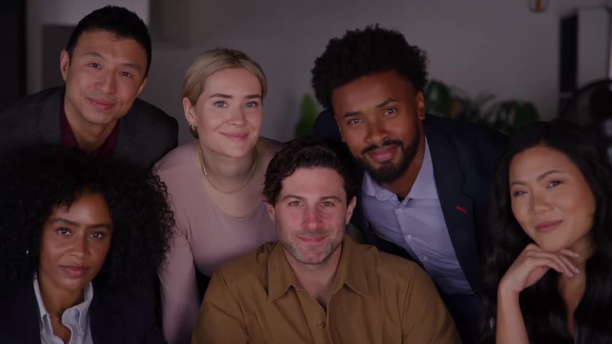 A diverse group of six coworkers wearing professional and business casual attire look directly at the camera and smile together in a close medium shot
