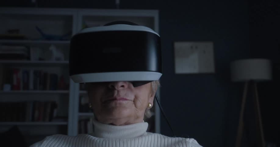 Woman wearing a white sweater sitting down on a yellow couch wearing a vr headset in a living room with flickering lights indoors during day time