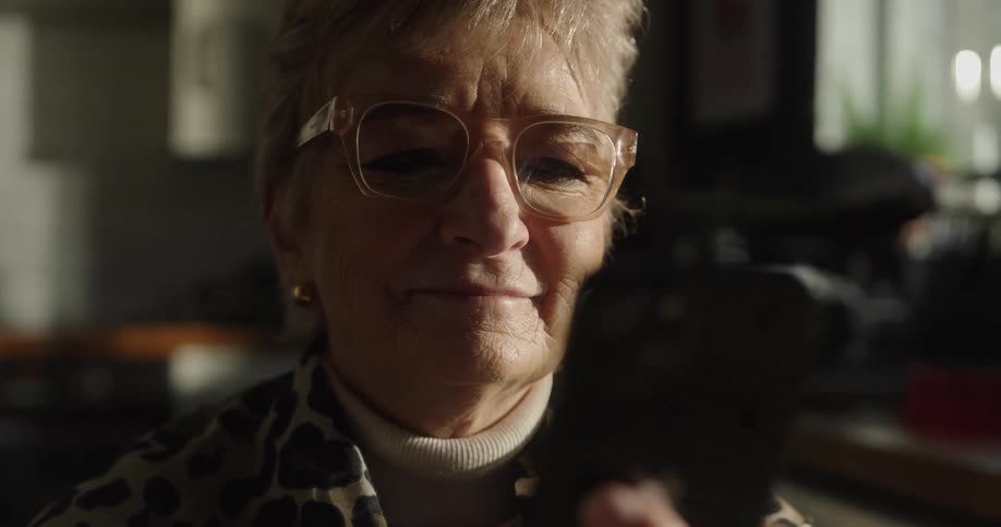 An older woman with glasses smiles warmly while looking at her phone in a sunlit room