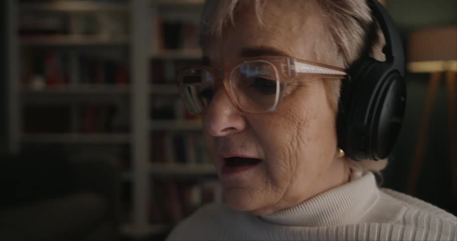 An elderly woman wears headphones and glasses while holding a smartphone immersed in content in a cozy book filled room