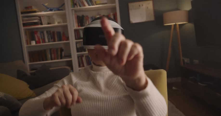 An older woman wearing a white turtleneck sweater and a virtual reality headset is seated in a yellow armchair actively moving her hands and arms to interact with the virtual environment and bookshelves and a modern floor lamp define the background