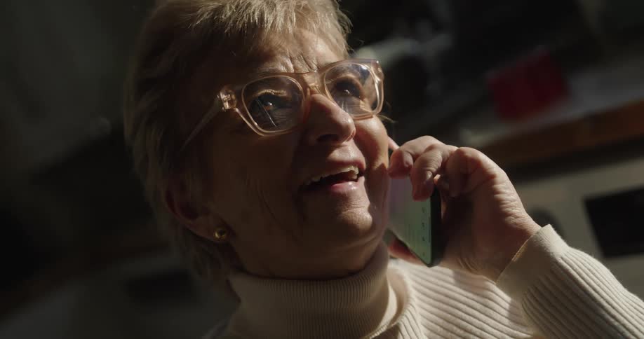 An elderly woman wearing glasses captures her happily talking on a smartphone with a bright smile on her face while strong light illuminates her from the side against a dark background
