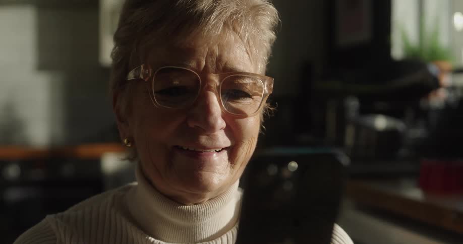 A closeup shot captures an elderly woman with glasses smiling and reacting joyfully as she looks at the screen of the smartphone held in her hand