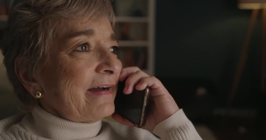 A close side view of an older woman with short gray hair and a white turtleneck talking animatedly on a smartphone in a dimly lit room