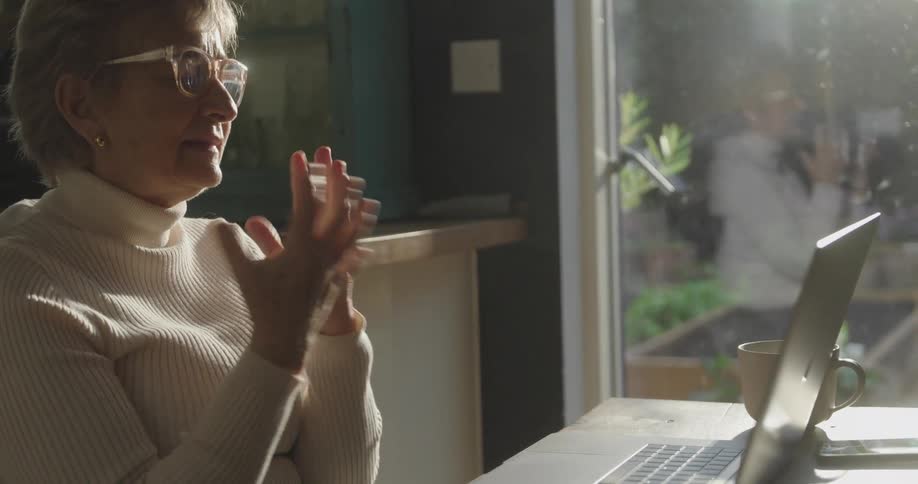 Adult woman wearing glasses sitting down while talking and gesticulating in front of a laptop on a table indoors during day time