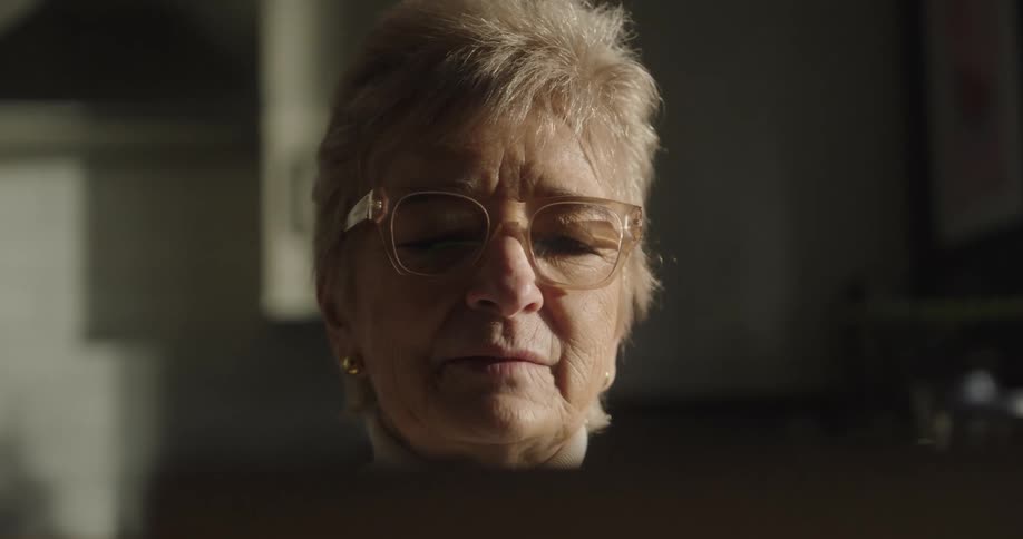 A close view of an older woman with short blonde hair and large glasses looking intently at a screen with dramatic light shining on her face