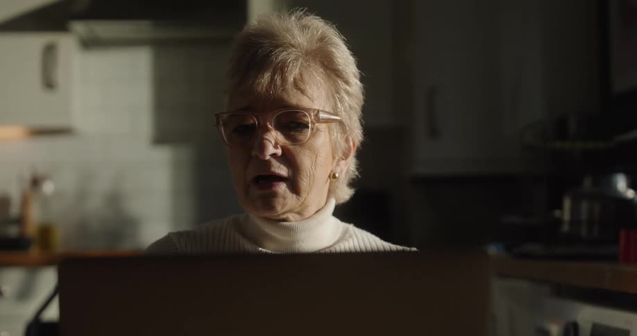 A closeup shot focuses on an elderly woman with glasses wearing a white turtleneck sitting in a dimly lit kitchen and looking intently at a laptop screen while reacting with a concerned expression