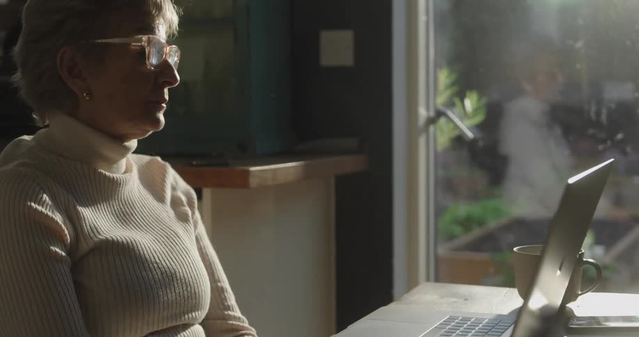 Adult woman wearing glasses sitting on a chair talking and gesticulating in front of a laptop computer on a table in front of her indoors during day time