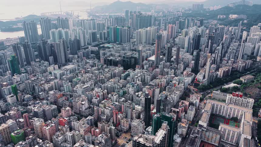 An expansive aerial shot captures a large city showcasing countless tightly packed highrise residential and commercial buildings extending across a valley toward distant hazy mountains and a large body of water crossed by a suspension bridge
