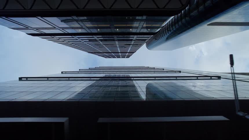 A lowangle view looks straight up between two tall modern glass skyscrapers against a light blue cloudy sky showing an urban environment