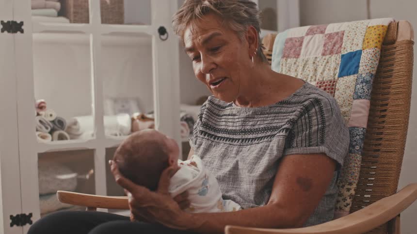 A close view of a grandmother sitting in a wooden rocking chair with a colorful quilt draped over it smiling and affectionately holding a newborn baby in a brightly lit room