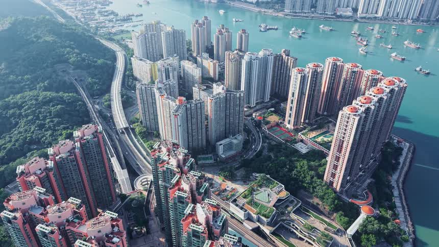 An aerial view shows densely packed tall residential and commercial buildings built on the green slope of a peninsula next to a bay filled with boats