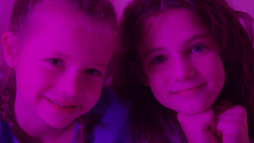 Two young girls are shown in a closeup shot illuminated by intense magenta and purple light giving the scene a vibrant stylized look they are smiling at the camera and then turn to share a secret or a private moment with each other